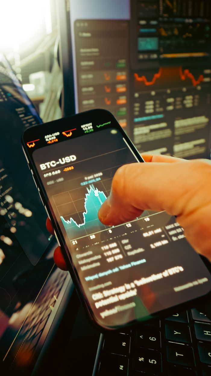 about-us-02 A hand holds a smartphone displaying BTC-USD chart amidst digital trading screens, highlighting crypto finance.