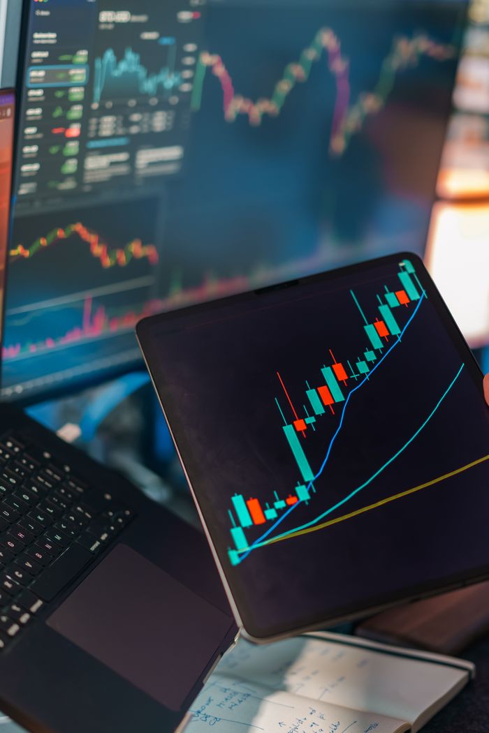 services-04 A tablet displaying a candlestick chart with a laptop screen showing stock market data for financial analysis.