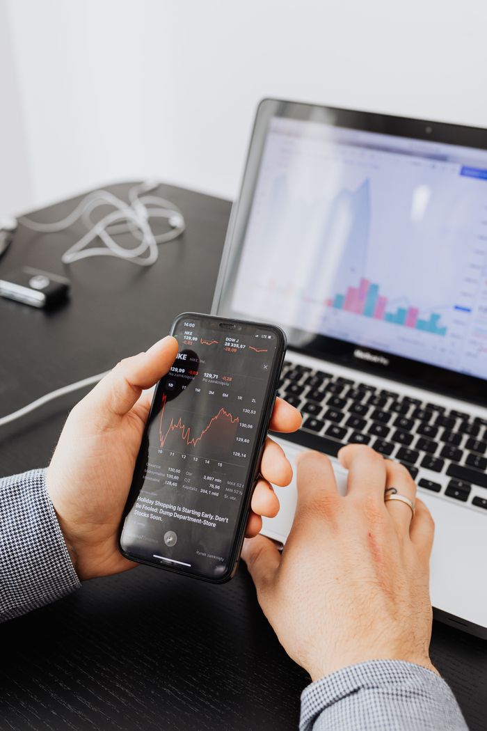 about-us-01 Hands holding smartphone analyzing stock market data next to laptop displaying charts.